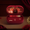 A red oval jewelry box shown open on a table, containing gold rings and a small gold pendant, with a red-and-gold applicator tool inside the lid. The scene is styled with dark red drapery and a gold decorative mirror edge.