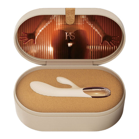 An open oval presentation case with a beige exterior and tan foam insert, containing a white curved personal massager with a transparent end piece. The inside lid features a warm orange-toned graphic with the letters 'KS'.