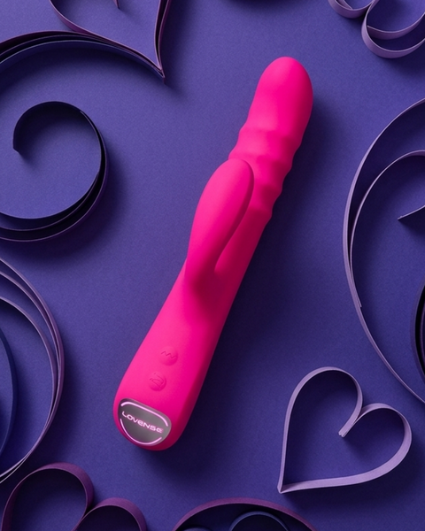 A bright pink personal massager with a textured shaft and a silver button, placed diagonally on a purple background decorated with layered paper hearts and swirls.