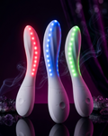 Three white handheld LED light therapy wand devices standing upright on a reflective black surface, each illuminated with a different color: red, blue, and green. Purple smoke effects and dark draped fabric are visible in the background.