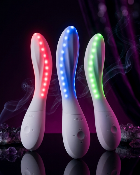 Three white handheld LED light therapy wand devices standing upright on a reflective black surface, each illuminated with a different color: red, blue, and green. Purple smoke effects and dark draped fabric are visible in the background.
